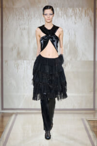 Model walking runway in an all-black avant-garde outfit: a cut-out glossy top and tiered fringed skirt with sheer gloves and heeled boots.