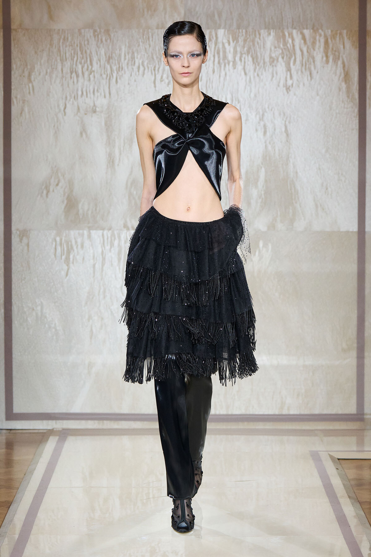 Model walking runway in an all-black avant-garde outfit: a cut-out glossy top and tiered fringed skirt with sheer gloves and heeled boots.