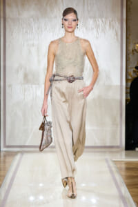 Model on a runway wearing a champagne sequin sleeveless top tucked into wide beige trousers, with a metallic rope belt and a small sparkling handbag.