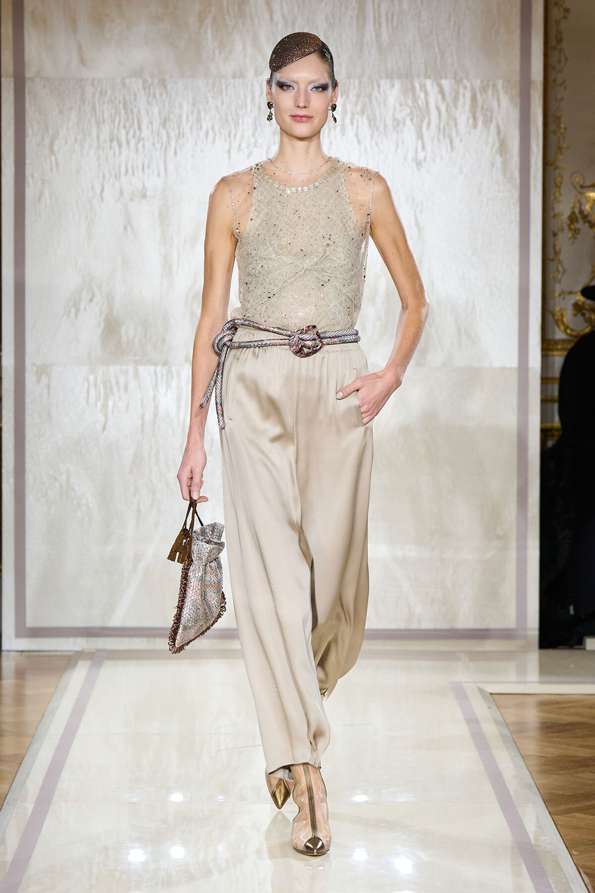 Model on a runway wearing a champagne sequin sleeveless top tucked into wide beige trousers, with a metallic rope belt and a small sparkling handbag.
