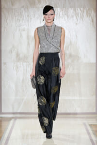 Model walking a runway in a silver sequined wrap top and black satin trousers with gold circular medallion prints, holding a textured silver clutch.