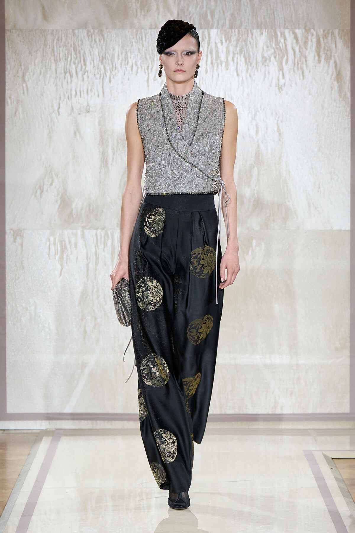 Model walking a runway in a silver sequined wrap top and black satin trousers with gold circular medallion prints, holding a textured silver clutch.