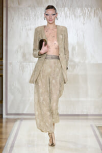 Runway model in a beige lace pantsuit with a sheer embellished top, holding a small clutch.
