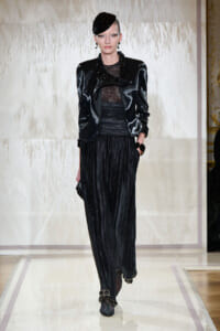 Model walking a runway in a black metallic blazer, sheer top, and wide-pleated black pants, with a sculpted black headpiece and statement jewelry.