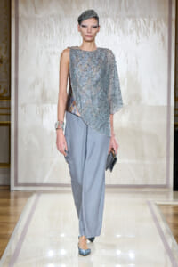 Runway model wearing a shimmering silver beaded top with pale blue wide-leg pants, holding a small clutch on the catwalk.”] }{