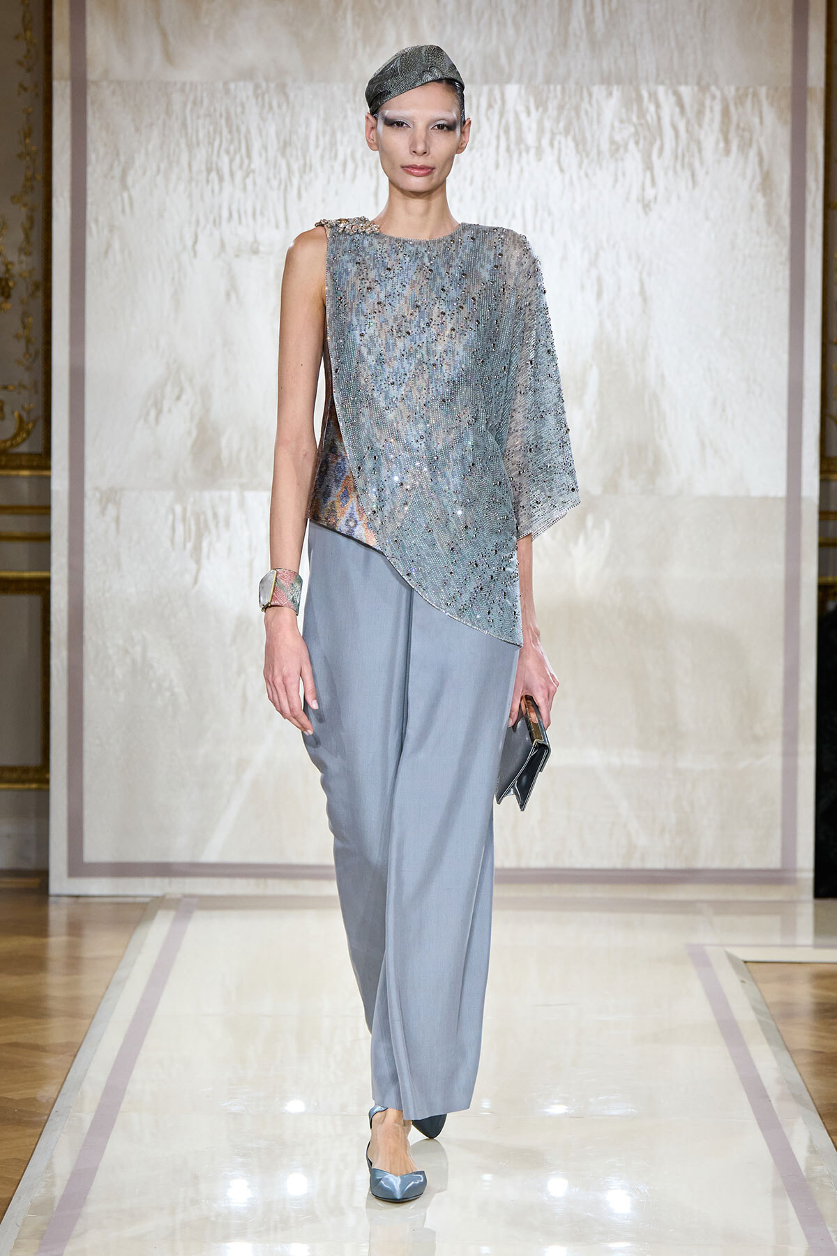Runway model wearing a shimmering silver beaded top with pale blue wide-leg pants, holding a small clutch on the catwalk.”] }{