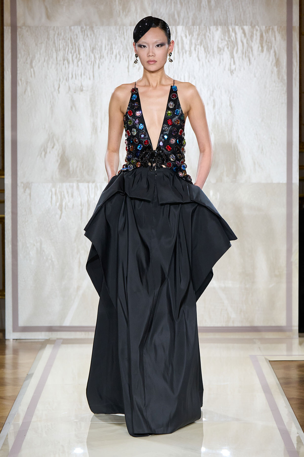 Model walking a runway in a black gown with a jewel-studded plunging bodice and a voluminous skirt.