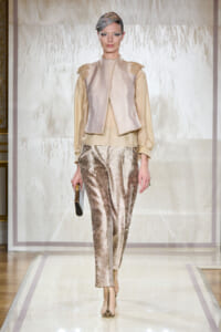 Model walking a runway in a champagne blouse with a metallic vest, silk-pattern pants, gold heels, a small gold clutch, and a metallic headpiece.
