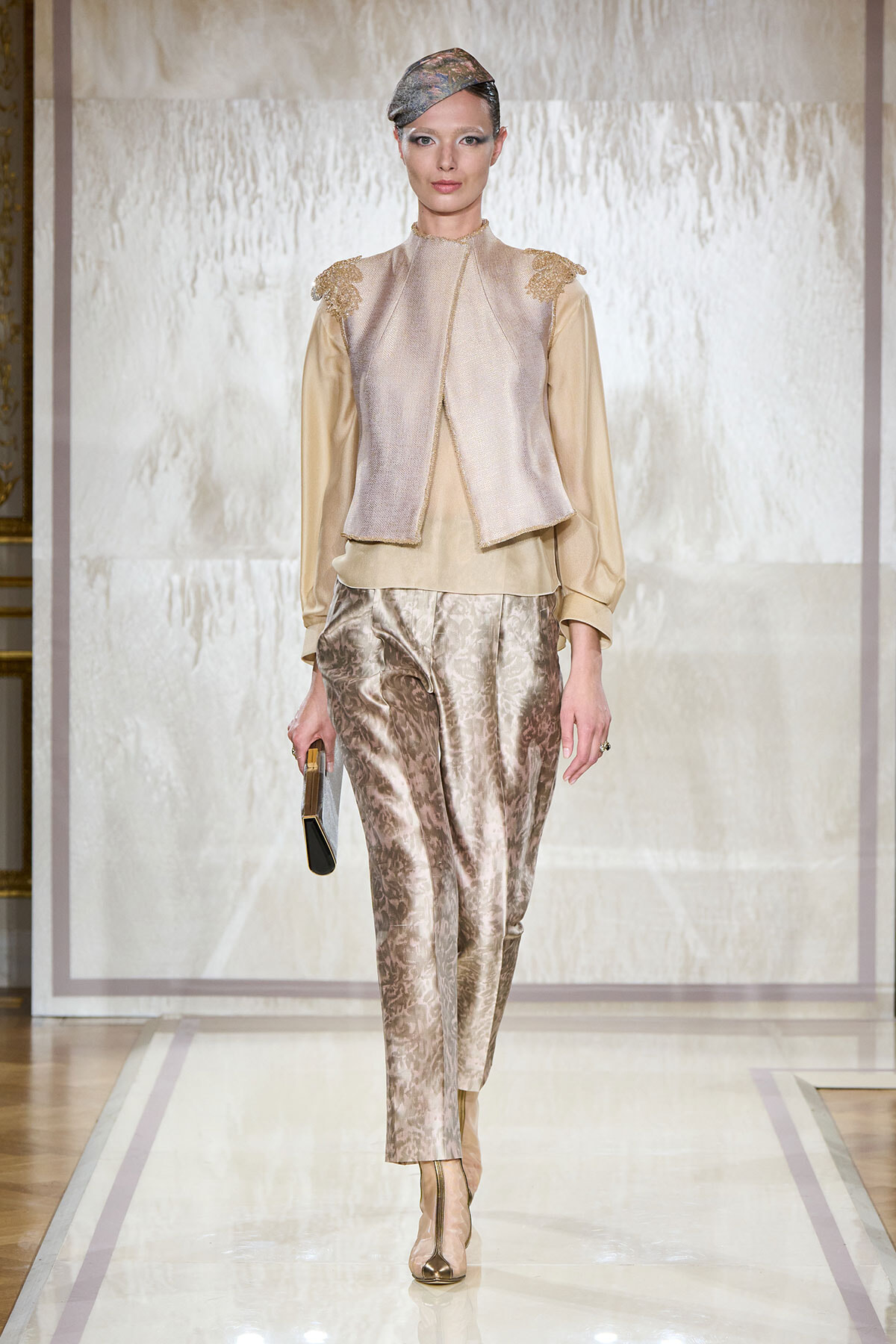 Model walking a runway in a champagne blouse with a metallic vest, silk-pattern pants, gold heels, a small gold clutch, and a metallic headpiece.