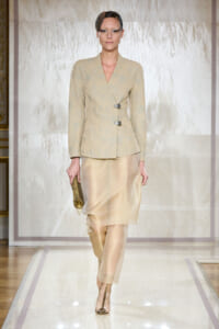 Model on a fashion runway wearing a beige, structured wrap blazer with metallic clasps and a matching satin skirt, holding a gold clutch with a small hat accessory.