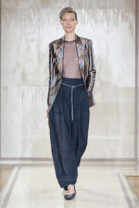 Model walking a runway in a metallic patterned blazer over a sheer taupe top and wide navy pants with a beaded belt and headband accessory.