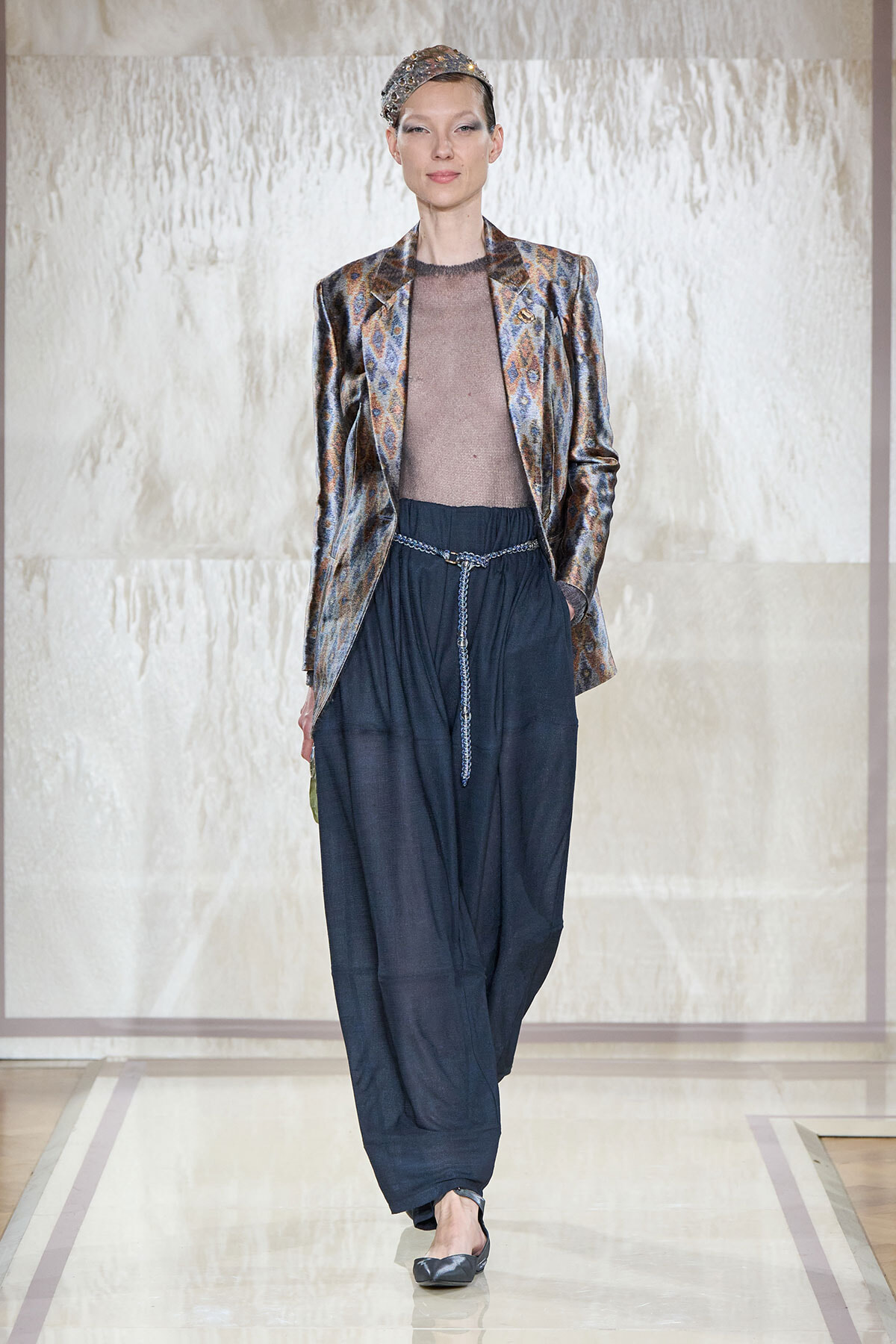 Model walking a runway in a metallic patterned blazer over a sheer taupe top and wide navy pants with a beaded belt and headband accessory.
