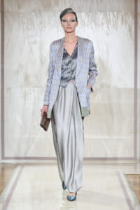 Model on a runway wearing a pale blue patterned blazer over a metallic vest and wide gray satin trousers, holding a textured clutch.