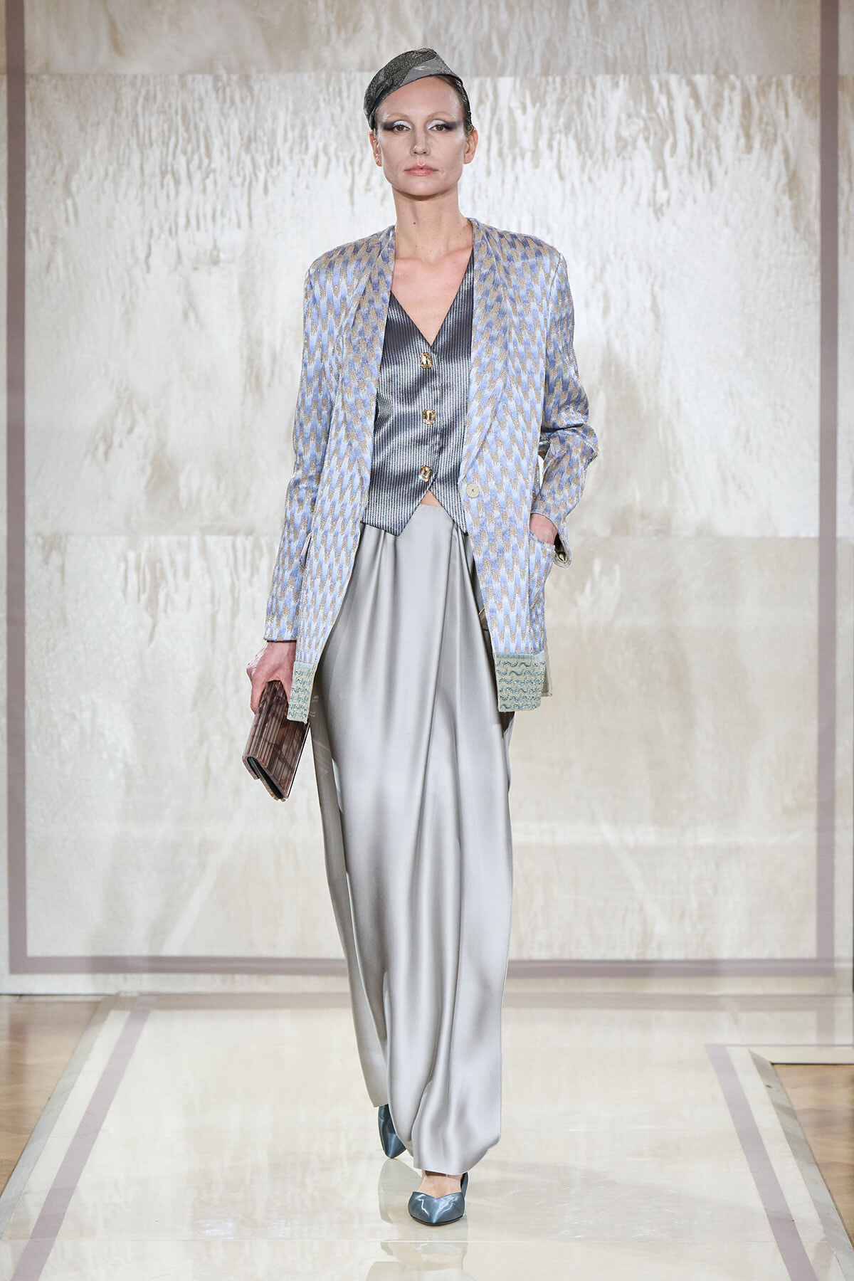 Model on a runway wearing a pale blue patterned blazer over a metallic vest and wide gray satin trousers, holding a textured clutch.