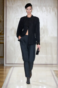 Runway model in an all-black ensemble: textured blouse, layered beaded necklace, voluminous pants, ankle boots, and a black cap, posing on a catwalk with a clutch