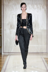 Model on a runway wearing a black velvet jacket with metallic trim over a black sequined crop top, black tailored pants, and black ankle boots, carrying a small black clutch with a chain strap.