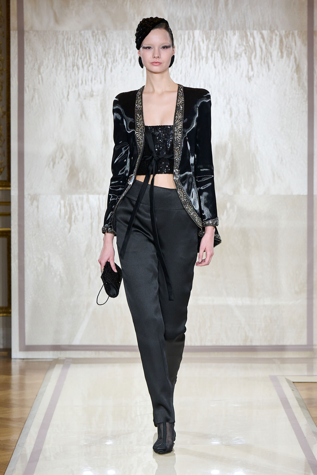 Model on a runway wearing a black velvet jacket with metallic trim over a black sequined crop top, black tailored pants, and black ankle boots, carrying a small black clutch with a chain strap.