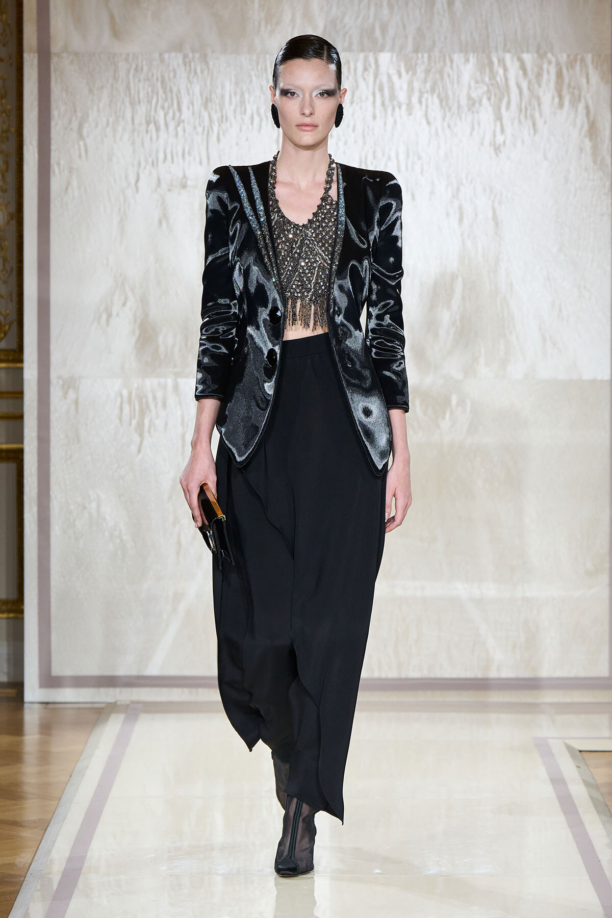 Model on a runway wearing a black velvet blazer, metallic beaded crop top, wide-leg black pants, ankle boots, and a small clutch.
