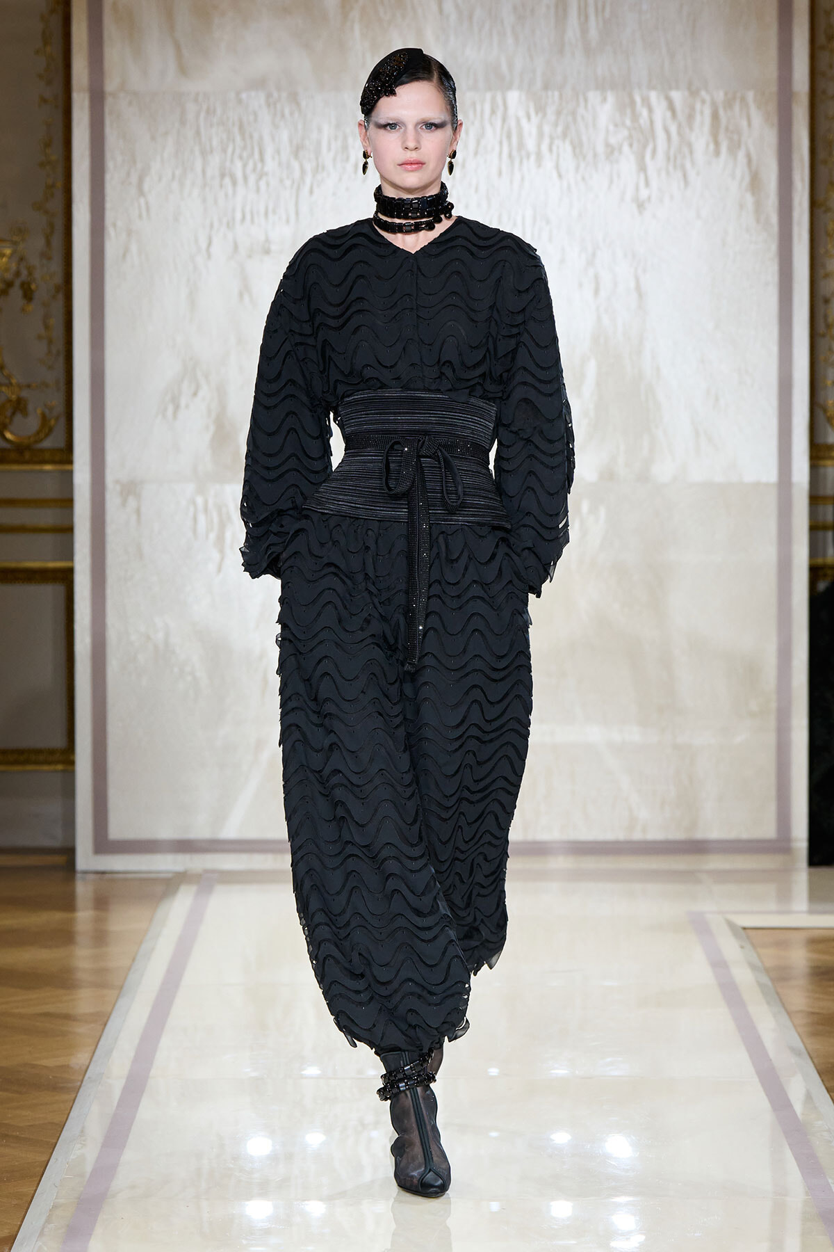 Model walks a black wavy-textured dress with a wide belt and choker, on a glossy runway backdrop.
