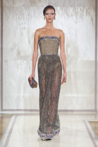 Model walking the runway in a strapless, silver beaded mesh gown with purple trim, holding a beaded clutch.