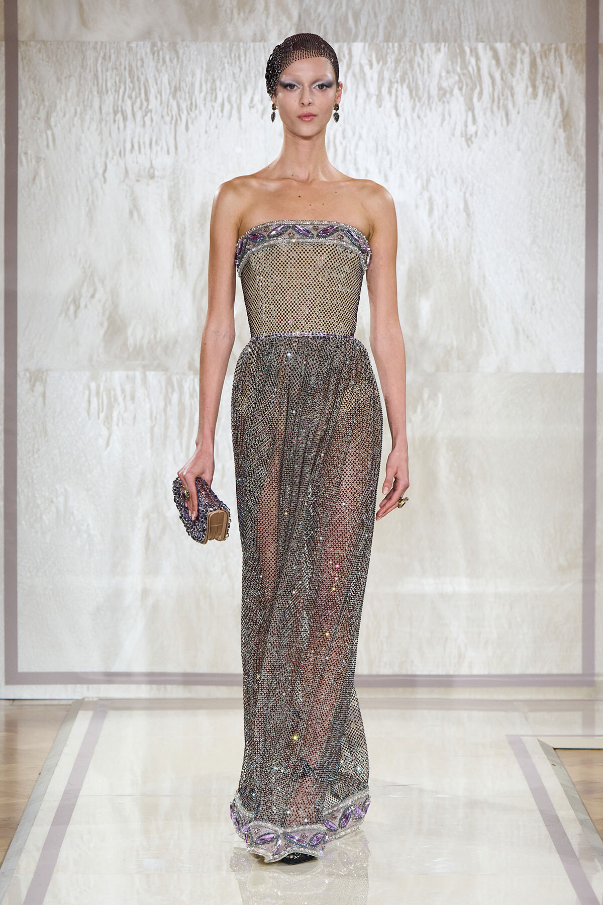 Model walking the runway in a strapless, silver beaded mesh gown with purple trim, holding a beaded clutch.