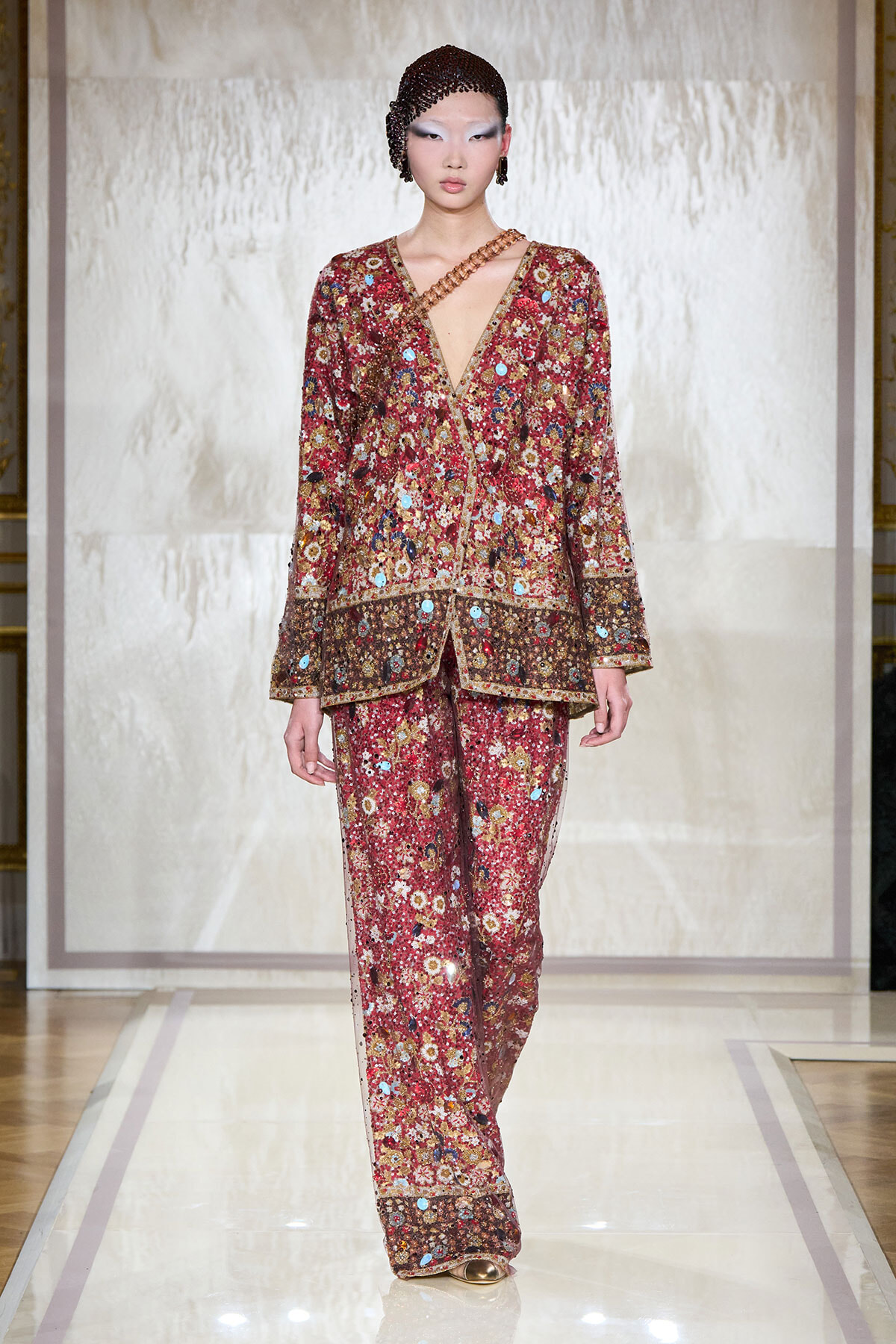 Runway model wearing a red floral embellished suit with a deep V neckline and gold trim.