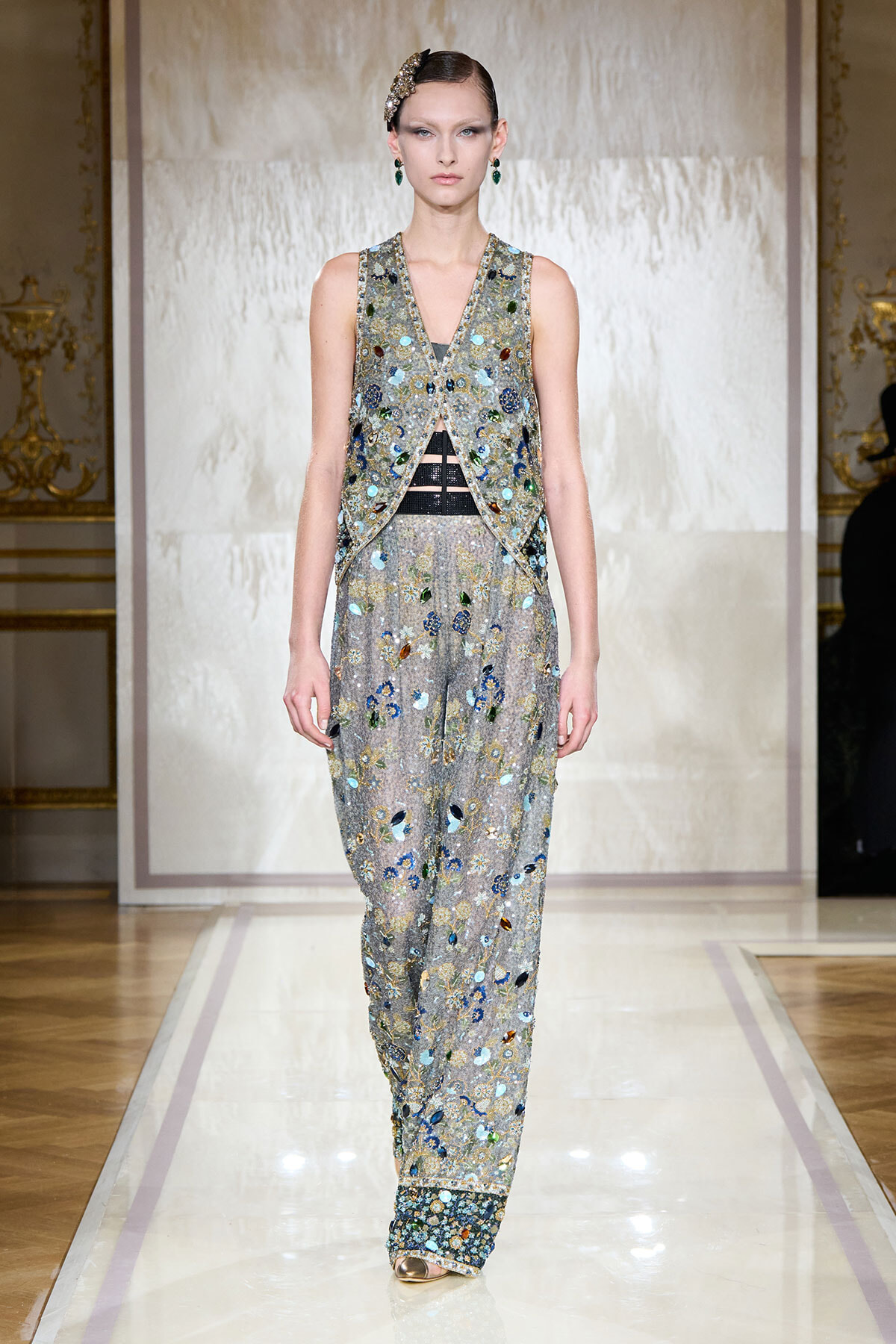 Model walking the runway in a silver, beaded sleeveless vest over a sheer, floral-embroidered jumpsuit with wide legs and jeweled cuffs; ornate headpiece.