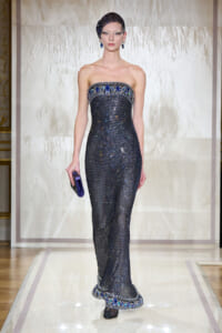 Model wearing a strapless, navy sequined gown with blue jeweled trim and matching headpiece on a runway.