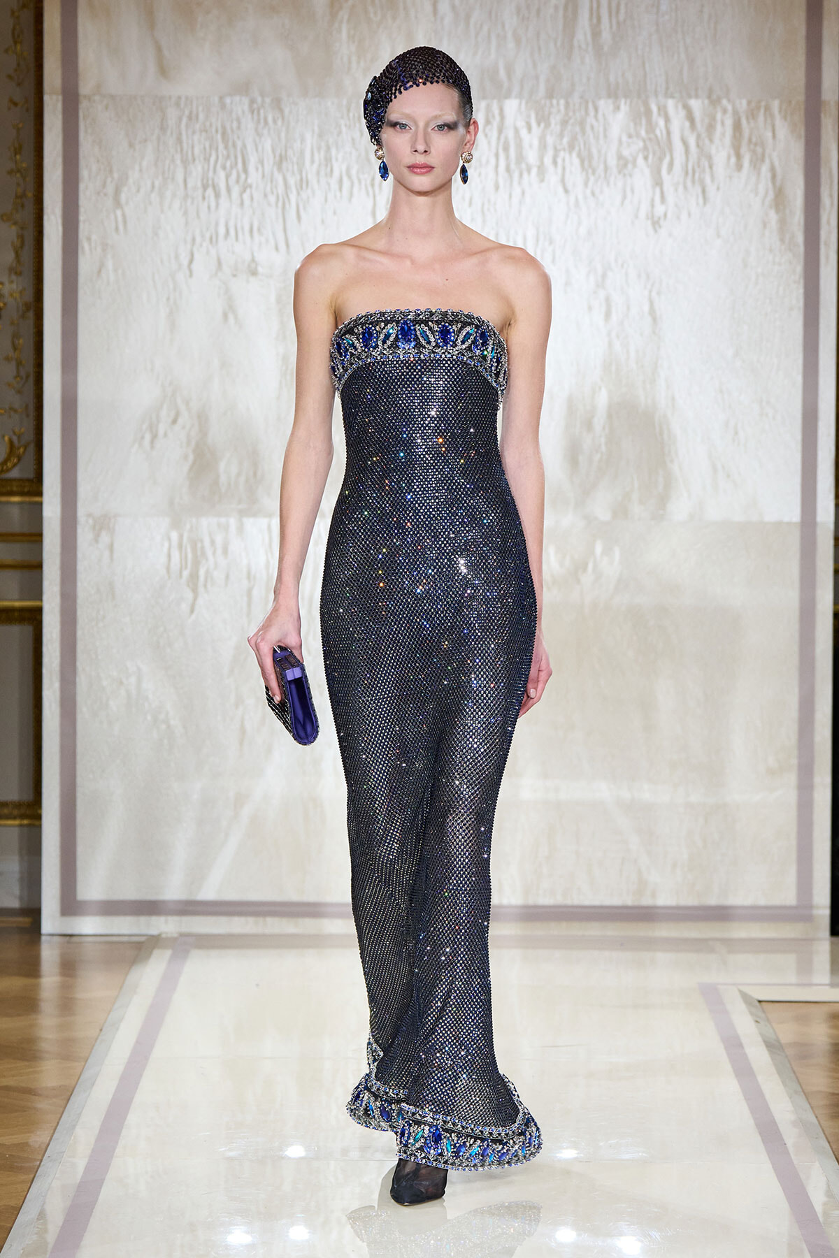 Model wearing a strapless, navy sequined gown with blue jeweled trim and matching headpiece on a runway.