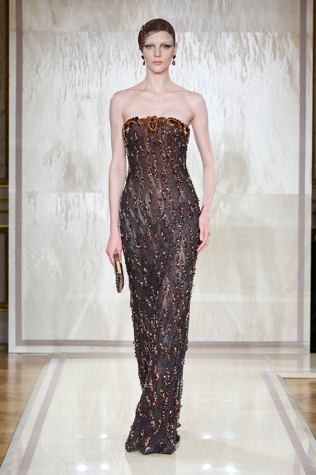 Model on a runway wearing a strapless, floor-length gown covered in dark beads and sequins, holding a small clutch.