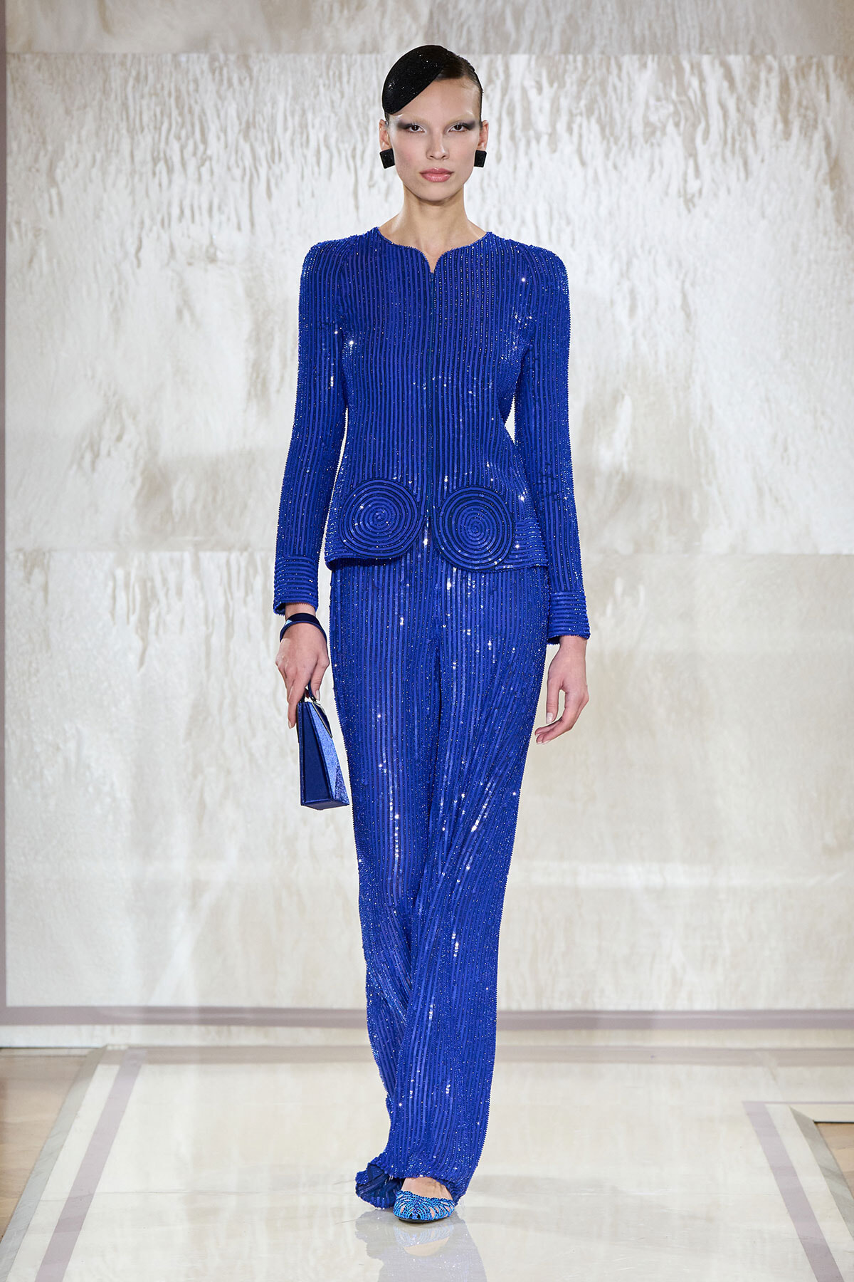 Model walking the runway in a royal blue sequined two-piece suit with circular swirl embellishments at the hips, holding a matching blue clutch.
