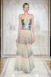 Model on a runway wearing a strapless metallic striped bodice with a layered, beaded tulle skirt and a green floral hair accessory.