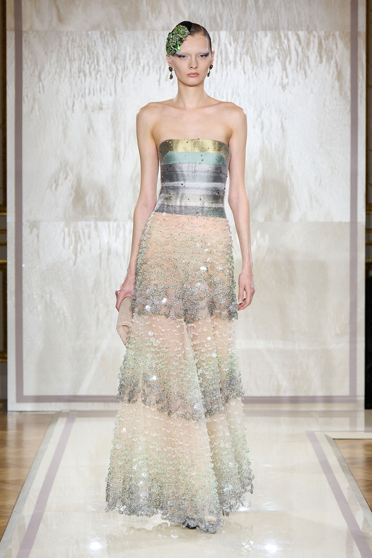 Model on a runway wearing a strapless metallic striped bodice with a layered, beaded tulle skirt and a green floral hair accessory.