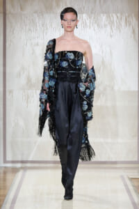 Model walking the runway in a black strapless top with blue floral embroidery, paired with satin pants and a fringed sheer cape.
