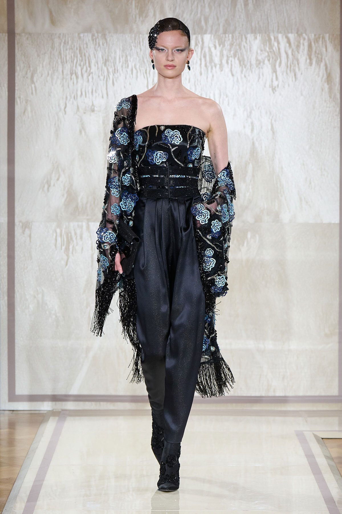 Model walking the runway in a black strapless top with blue floral embroidery, paired with satin pants and a fringed sheer cape.