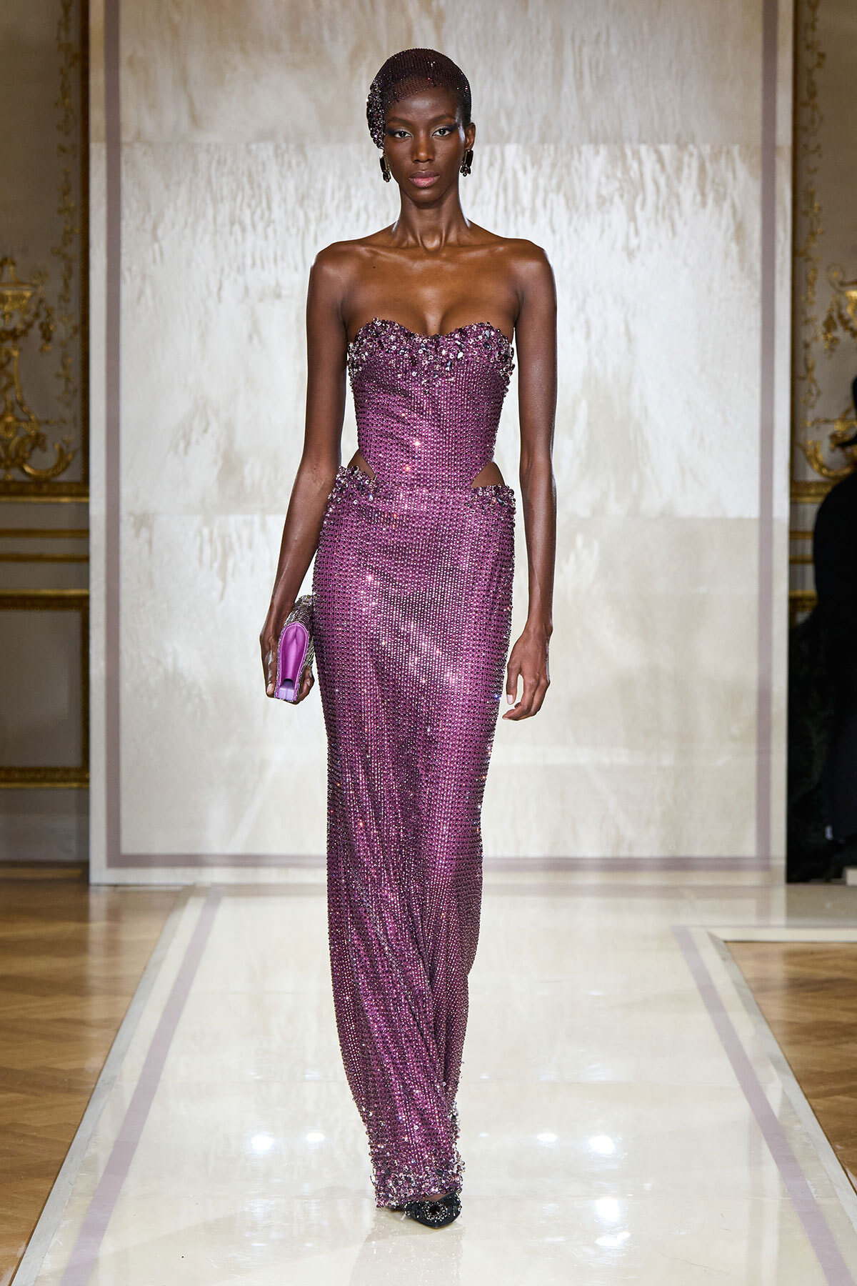 Model walks the runway in a strapless, floor-length purple-sequined gown with cutouts at the waist, holding a matching purple clutch.