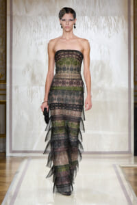 Model on a fashion runway wearing a strapless, multi-layered lace gown with dark, metallic tones and a jeweled headpiece.