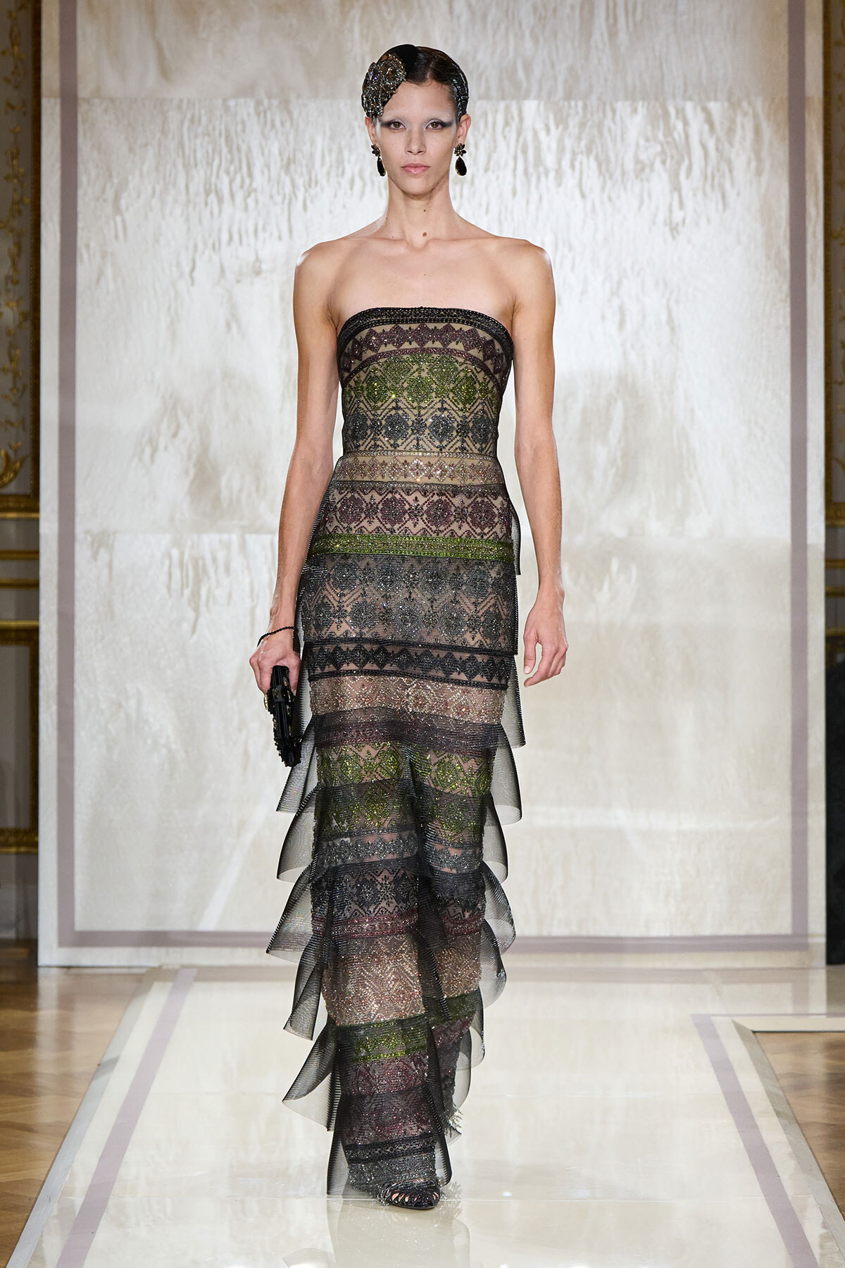 Model on a fashion runway wearing a strapless, multi-layered lace gown with dark, metallic tones and a jeweled headpiece.