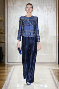 Runway model wearing a cobalt blue embellished jacket and wide-leg pleated navy pants, carrying a blue clutch.