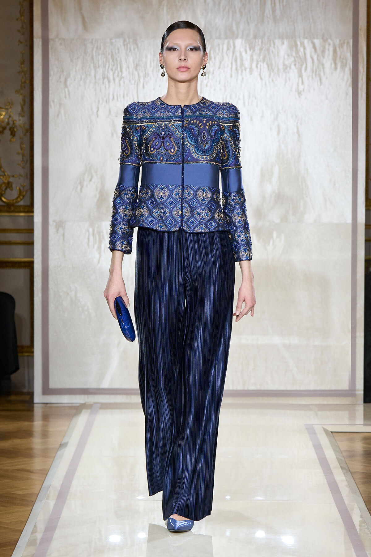 Runway model wearing a cobalt blue embellished jacket and wide-leg pleated navy pants, carrying a blue clutch.