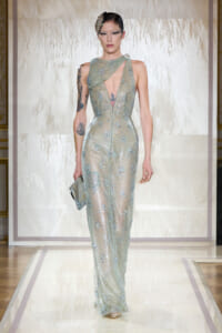 Model walks runway in a pale aqua sequined gown with a sheer plunging front, holding a silver clutch.