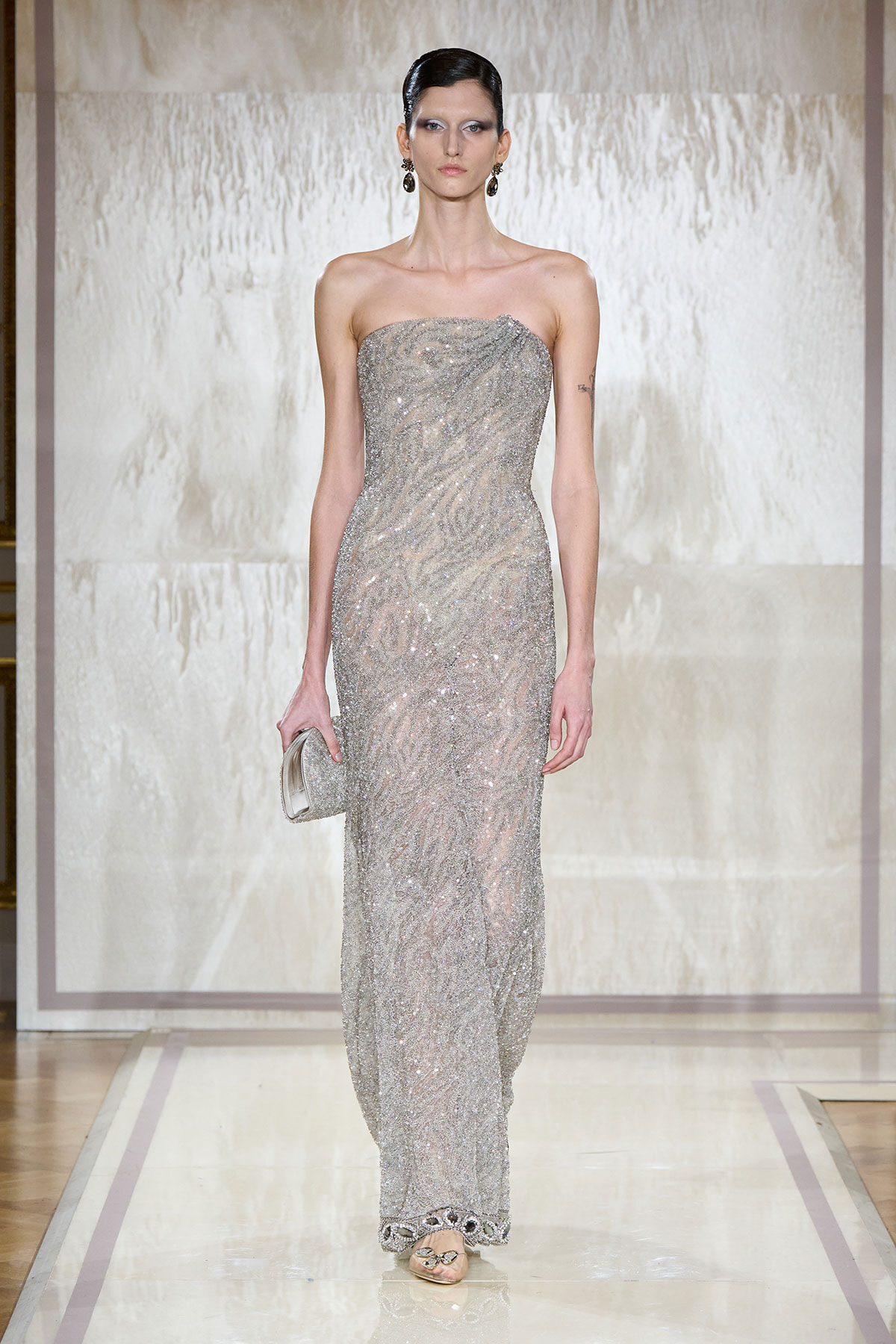 Model walks the runway in a strapless silver sequin gown, holding a small silver clutch with her hair pulled back.