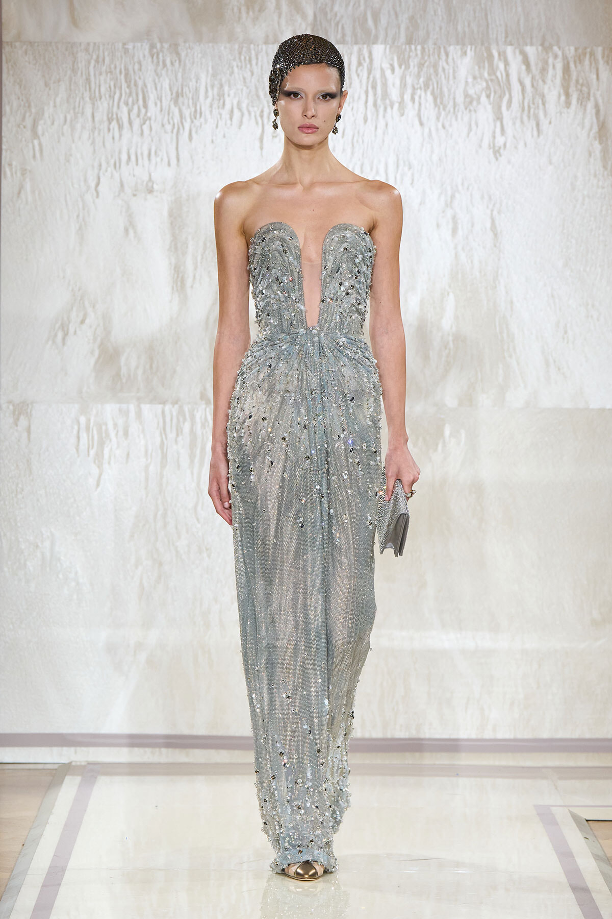 Model walking a runway in a silver, beaded, floor-length gown with a plunging V neckline and cinched waist, holding a small clutch.