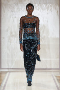 Model walking a runway in a sheer black dress densely embellished with blue beads and a matching black bag.