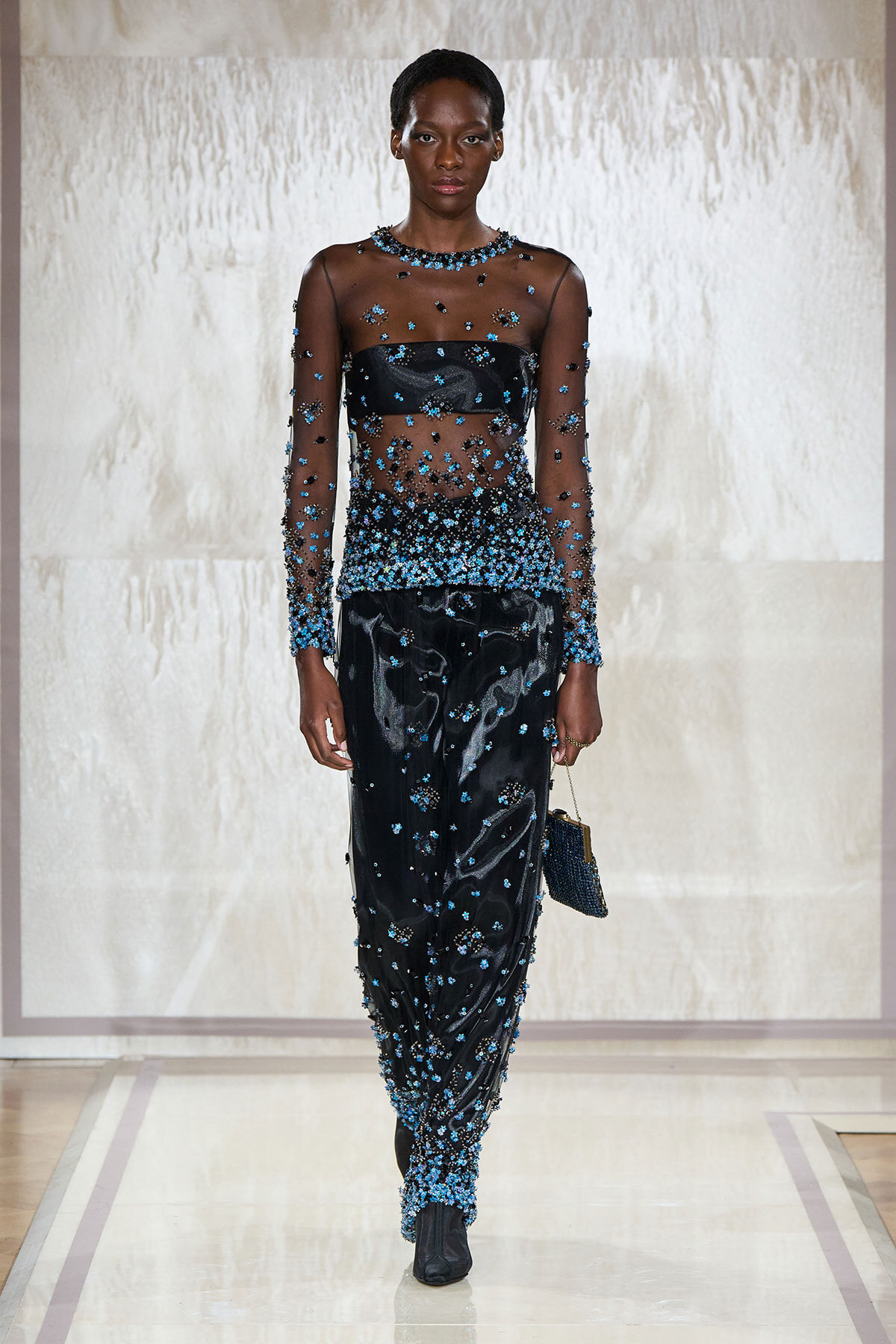 Model walking a runway in a sheer black dress densely embellished with blue beads and a matching black bag.