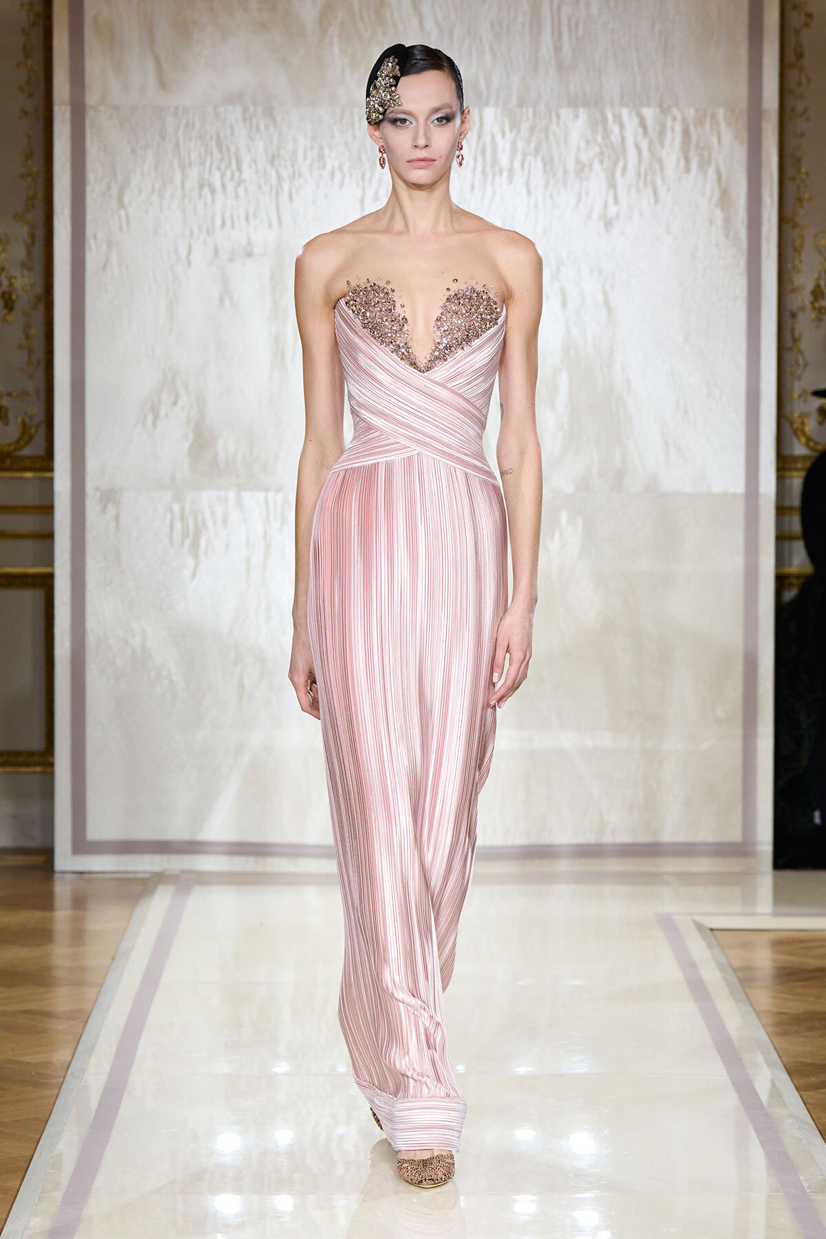 Model on a runway wearing a pale pink, pleated gown with a heart-shaped beaded bust and a decorative headpiece.