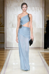 Model on a runway wearing a pale blue one-shoulder gown with draped waist, crystal belt, and black clutch.