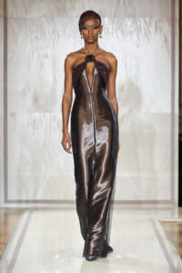 Model walking the runway in a glossy, metallic gown with a maroon halter neckline and central slit, posing elegantly.