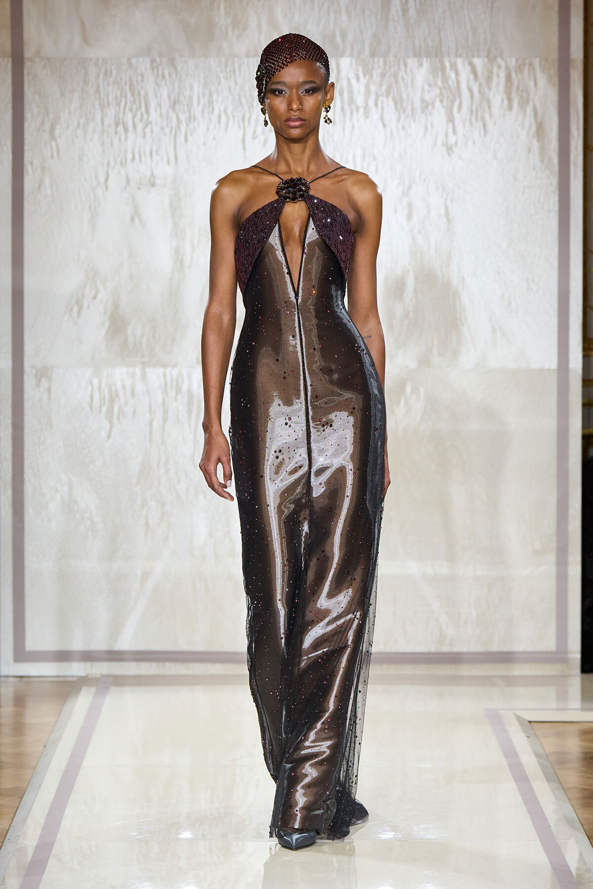 Model walking the runway in a glossy, metallic gown with a maroon halter neckline and central slit, posing elegantly.