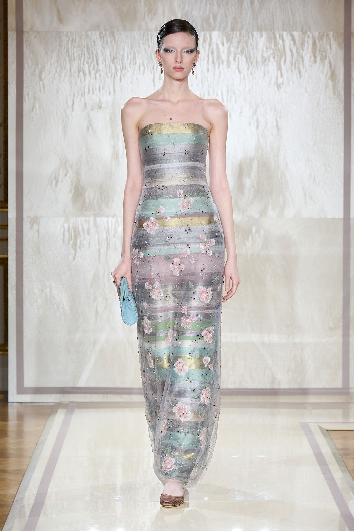 Strapless pastel striped gown with pink flowers on a model walking a runway, holding a light blue clutch.